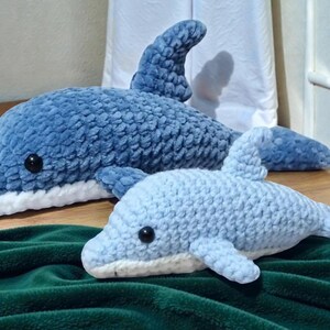 Peut inclure: Deux dauphins en peluche crochetés, dans des tons de bleu. Le plus grand est bleu foncé, le plus petit bleu clair. Les deux ont des yeux noirs et un ventre blanc. Ils sont posés sur une surface en bois et une couverture verte.