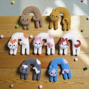 May include: A collection of handmade crochet cat toys in various colors, including brown, yellow, white with orange spots, white with pink spots, white with red spots, gray with white spots, and blue with purple spots. The toys are arranged on a wooden surface.