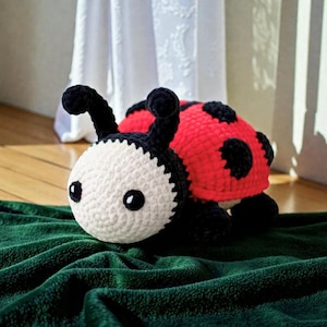 May include: A handmade crochet ladybug plush toy. The ladybug features a white head with black eyes and antennae, a red body with black spots, and black legs. It is resting on a green blanket.