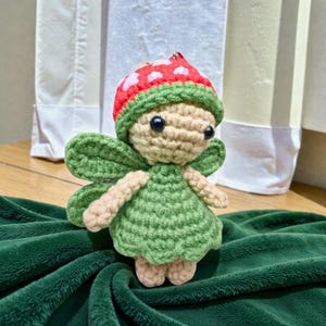 May include: A handmade crochet fairy doll with a mushroom cap hat. The fairy has green wings and a green dress. The hat is red with white spots. The doll is beige with black eyes and is sitting on a green blanket.