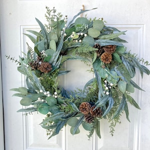 Op de afbeelding: Een groene en bruine krans gemaakt van eucalyptusbladeren, dennenappels en witte bessen. De krans is perfect voor een feestelijke of winterse decoratie.