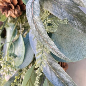 Op de afbeelding: Close-up van een eucalyptuskrans met een dennenappel en groene bladeren. De bladeren zijn lichtgroen van kleur en hebben een zachte, fluweelzachte textuur.