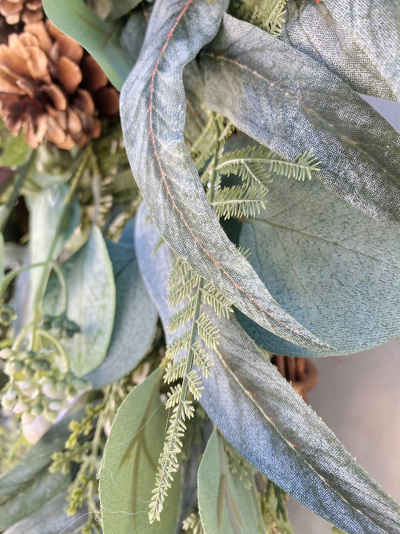 Op de afbeelding: Close-up van een eucalyptuskrans met een dennenappel en groene bladeren. De bladeren zijn lichtgroen van kleur en hebben een zachte, fluweelzachte textuur.