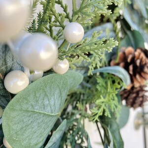 Op de afbeelding: Een close-up van een groene en witte kerstkrans met dennenappels, eucalyptusbladeren en witte bessen.