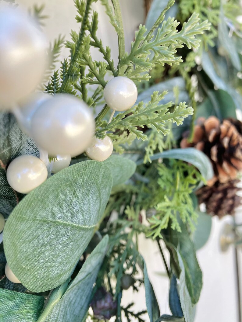 Op de afbeelding: Een close-up van een groene en witte kerstkrans met dennenappels, eucalyptusbladeren en witte bessen.