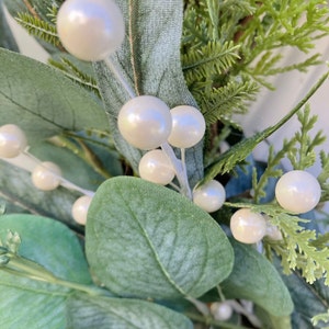 Op de afbeelding: Een close-up van een eucalyptuskrans met witte parel-achtige bessen. De krans is gemaakt van kunstgroen en is perfect om elke woning een vleugje elegantie te geven.