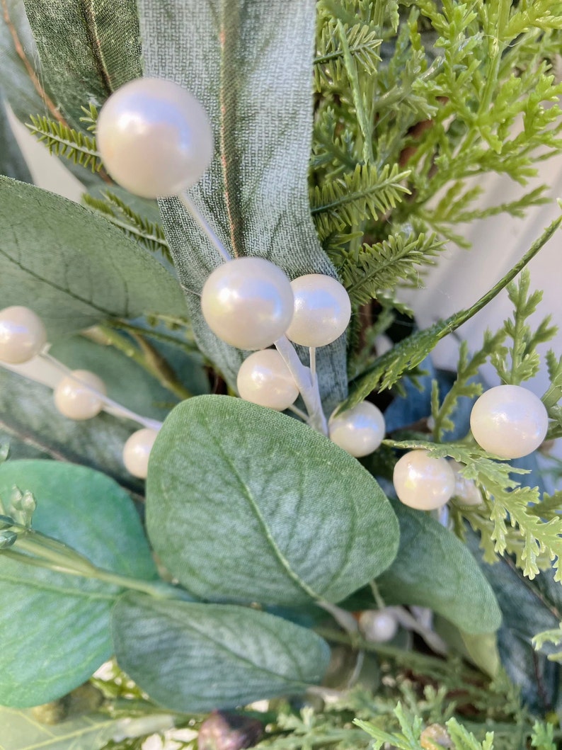 Op de afbeelding: Een close-up van een eucalyptuskrans met witte parel-achtige bessen. De krans is gemaakt van kunstgroen en is perfect om elke woning een vleugje elegantie te geven.