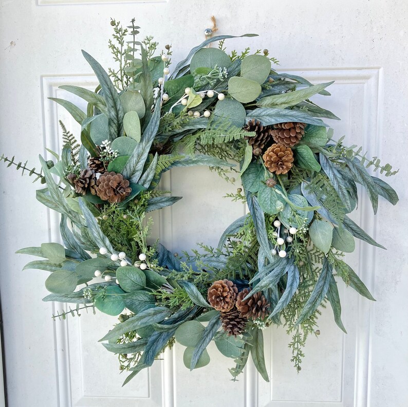 Op de afbeelding: Een groene en witte eucalyptuskrans met bruine dennenappels en witte bessen.