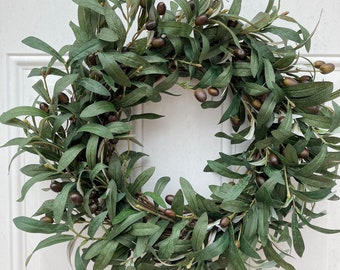 May include: A green olive branch wreath with brown olives. The wreath is hanging on a white door.