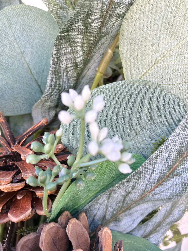 Op de afbeelding: Een close-up van een eucalyptuskrans met witte bloemen en dennenappels. De krans is gemaakt van kunstmatige eucalyptusbladeren en heeft een rustieke, natuurlijke uitstraling.