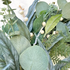 Op de afbeelding: Close-up van een groene eucalyptuskrans met witte parelmoerachtige bessen. De krans is gemaakt van kunstgroen en is perfect om een ​​natuurlijke touch aan je interieur te geven.