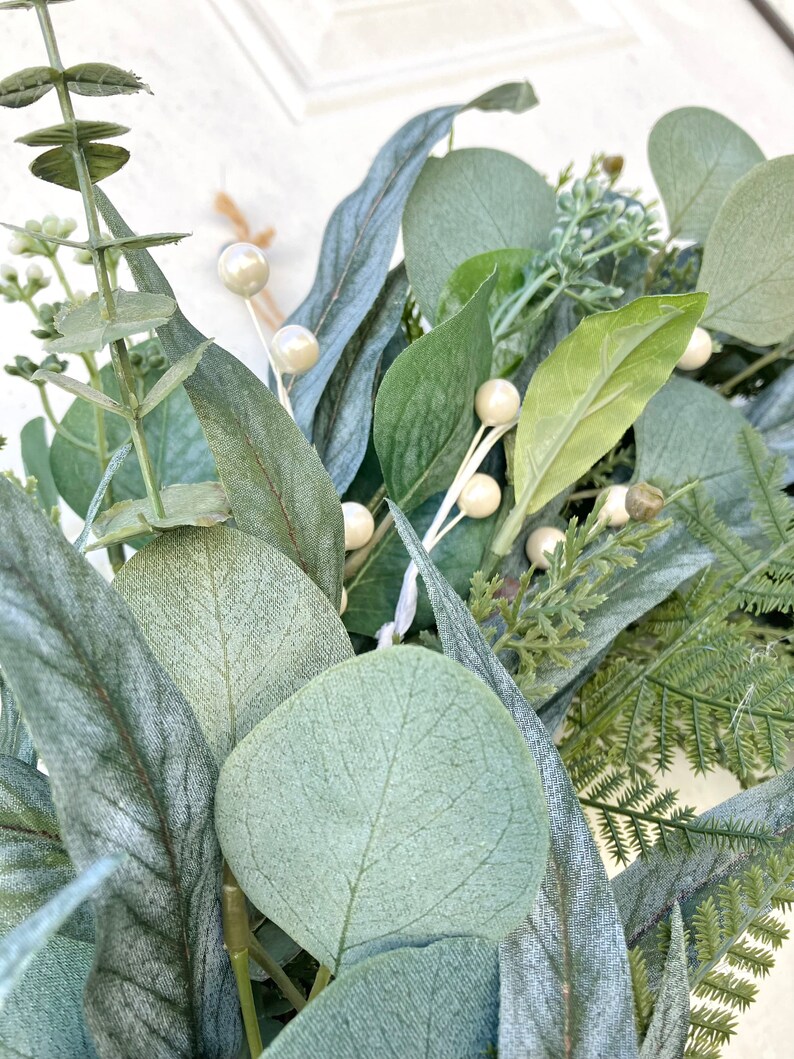 Op de afbeelding: Close-up van een groene eucalyptuskrans met witte parelmoerachtige bessen. De krans is gemaakt van kunstgroen en is perfect om een ​​natuurlijke touch aan je interieur te geven.