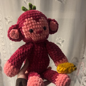 May include: A handmade crochet monkey doll in shades of red and pink, with a green leafy top and black eyes. The monkey is holding a small yellow banana. The plush toy is soft and cuddly.