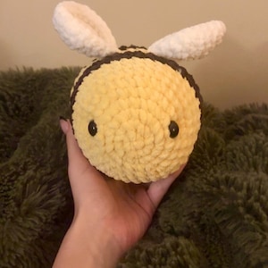 May include: A handmade, plush bee toy with a yellow body, black stripes, and white wings. The bee has black button eyes and is held in a hand. The toy is made of soft, textured yarn.