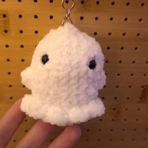 Könnte beinhalten: Ein weißer, flauschiger Geister-Schlüsselanhänger mit schwarzen Knopfaugen. Die Plüschfigur ist an einem silbernen Schlüsselring befestigt. Der Schlüsselanhänger besteht aus einem weichen, strukturierten Material und ist ein niedliches, skurriles Accessoire.