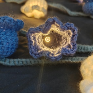 May include: Close-up of a string of crochet lights. The lights are shaped like flowers and are in shades of blue and white. Each flower has a small light inside, creating a warm glow. The string is a natural beige color.