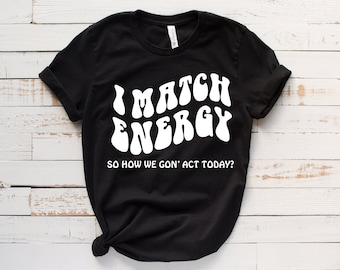 May include: Black t-shirt with white text that reads "I match energy so how we gon' act today?" The text is in a retro, wavy font.