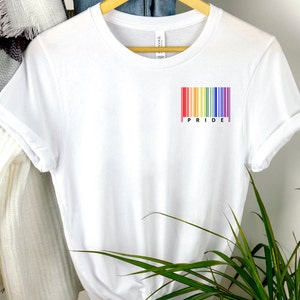 Op de afbeelding: Wit T-shirt met een regenboog-streepjescode en het woord "PRIDE" gedrukt op de voorkant.