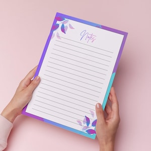 Puede incluir: Un bloc de notas rayado con un borde morado, azul y turquesa y detalles florales. La palabra "Notes" está escrita en cursiva en la parte superior. El bloc de notas está sostenido por dos manos sobre un fondo rosa.