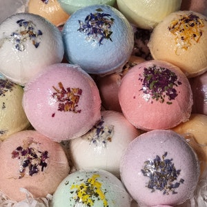May include: A selection of pastel-coloured bath bombs, including pink, blue, and green. Each round bath bomb is wrapped in clear plastic and decorated with dried flower petals. The bath bombs are arranged in white paper.