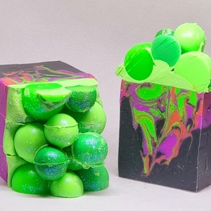 May include: Two bars of handmade soap with a black and purple swirl design. One bar is topped with green and black spheres, the other has a green and black swirl design.