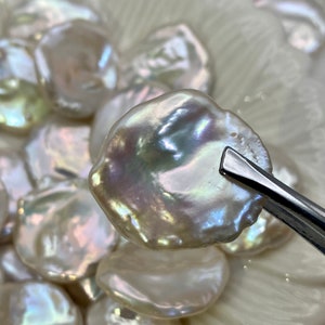 May include: A close-up of a large, iridescent white pearl with a slightly irregular shape. The pearl is being held by tweezers and is surrounded by other pearls of varying sizes.