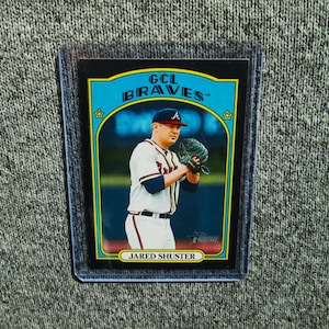 May include: A baseball card featuring Jared Shuster of the GCL Braves. The card has a blue and yellow border with the team name at the top. Shuster is wearing a Braves uniform and a baseball glove. The card is in a protective sleeve.