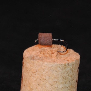 May include: A brown foam fly tied on a hook, sitting on a cork.