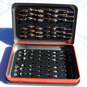 May include: Open metal tackle box with two compartments filled with various fishing flies. The flies are arranged neatly in rows, showcasing a variety of colors and designs. The box has a black interior and an orange trim.
