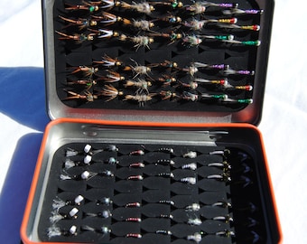 May include: Open metal tackle box with two compartments filled with various fishing flies. The flies are arranged neatly in rows, showcasing a variety of colors and designs. The box has a black interior and an orange trim.
