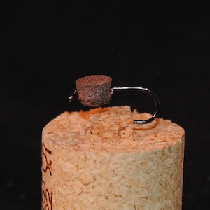 May include: A brown fly tying material is tied to a black fishing hook. The hook is sitting on a cork.
