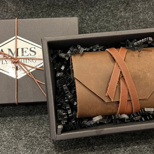 Może przedstawiać: Brązowy skórzany dziennik wędkarski z zapięciem na skórzany pasek, zapakowany w czarne pudełko z białą etykietą z napisem "AMES FLY FISHING".