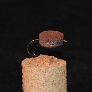 May include: A brown foam fly tied to a gold hook. The fly is sitting on a cork base.