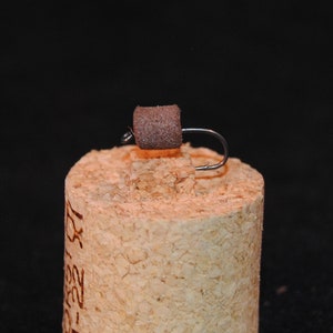 May include: A brown foam fly tied to a silver hook. The fly is sitting on a cork.