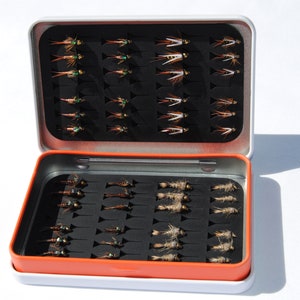 May include: A metal fly fishing box with two rows of compartments filled with various flies. The box is open and the flies are visible.