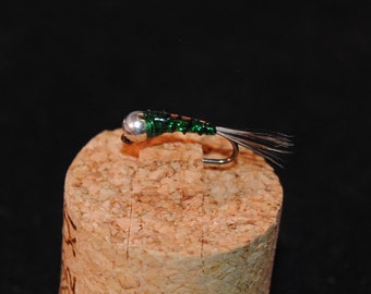 May include: A close-up of a fishing fly, resting on a cork. The fly has a silver head, a green body, and a curved hook. The fly's tail is made of thin, dark gray and white fibers. The background is black.