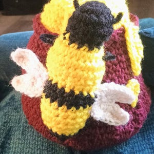 May include: A crocheted maroon pot with a yellow textured top and a large, three-dimensional crocheted bee attached to the front. The bee features a black head, a yellow and black striped body, and two white wings extending from its sides.