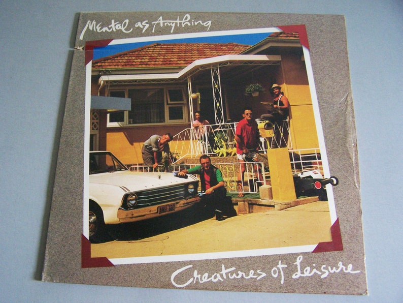以下が含まれることがあります： メンタル・アズ・エニシングの「Creatures of Leisure」のアルバムカバー。カバーには、家の前でグループの人々が写っている写真があり、白い車が前景にあります。アルバムタイトルとバンド名がカバーに書かれています。