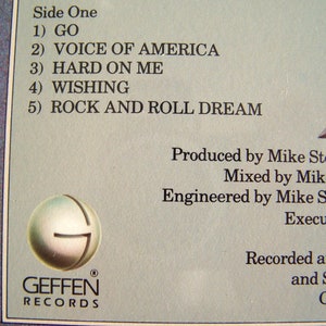 May include: Close-up of a record album's back cover, featuring a tracklist: "GO," "VOICE OF AMERICA," "HARD ON ME," "WISHING," and "ROCK AND ROLL DREAM." The Geffen Records logo is visible. The album credits include production, mixing, and engineering details.