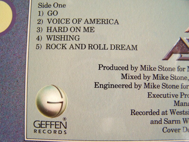 May include: Close-up of a record album's back cover, featuring a tracklist: "GO," "VOICE OF AMERICA," "HARD ON ME," "WISHING," and "ROCK AND ROLL DREAM." The Geffen Records logo is visible. The album credits include production, mixing, and engineering details.