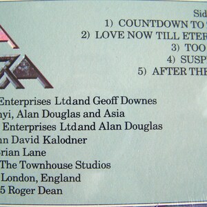 May include: Close-up of a vintage album cover with the band name "Asia" in a stylized font. The tracklist for Side Two is visible, including songs like "Countdown to Zero" and "Love Now Till Eternity." The album was released in 1985.