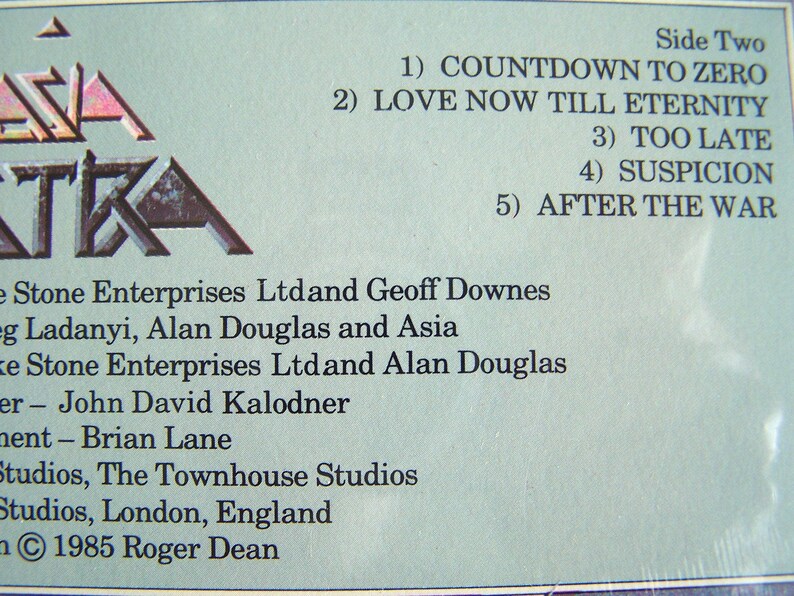 May include: Close-up of a vintage album cover with the band name "Asia" in a stylized font. The tracklist for Side Two is visible, including songs like "Countdown to Zero" and "Love Now Till Eternity." The album was released in 1985.
