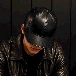 May include: A black baseball cap constructed from a smooth, semi-glossy material featuring a structured six-panel design with a curved brim, a central top button, and visible stitching along the seams of the crown and the edge of the visor.