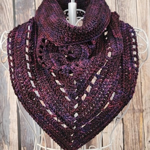 May include: A handmade, crocheted cowl scarf in shades of deep purple, burgundy, and pink. The scarf is displayed on a white mannequin, showcasing its textured stitch pattern and openwork design. The cowl is a triangular shape.