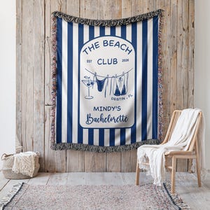 May include: A blue and white striped beach blanket with a decorative fringe. The blanket features the text "The Beach Club" and "Mindy's Bachelorette" with beach-themed illustrations. The blanket is hanging on a wooden wall.