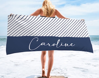 Puede incluir: Una toalla de playa blanca con rayas azules diagonales y una banda azul sólida en la parte inferior. El nombre "Caroline" está escrito en blanco en la banda azul.
