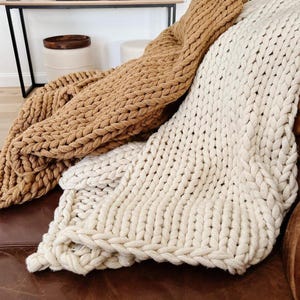 May include: Two chunky knit blankets in the image. One is a warm tan color, and the other is a creamy off-white. The blankets are draped over a brown leather couch, showcasing their thick, textured weave. The cozy, oversized design suggests comfort and warmth.