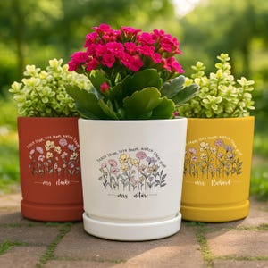 May include: Three colorful ceramic flower pots with plants. The pots are in shades of rust, white, and yellow. Each pot has a floral design and the text "teach them, love them, watch them grow". The names "Mrs. Clarke", "Mrs. Notar", and "Mrs. Richard" are also printed on the pots.