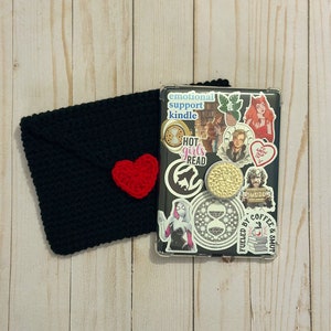 May include: A black crocheted envelope with a red heart on top, next to a clear phone case with various stickers. The stickers include a woman with red hair, a woman with a spider web design, a man with a beard, a coffee cup, and the text "emotional support kindle", "hot girls read", and "fueled by coffee & smut".