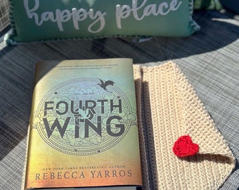 May include: A book titled "Fourth Wing" by Rebecca Yarros, a New York Times bestselling author, is on a beige crocheted book cover with a red heart. The book cover has a design of a bird flying in a circle with the words "Fly...or Die" above it. The book is on a gray surface with a green pillow that says "my happy place" in white lettering.